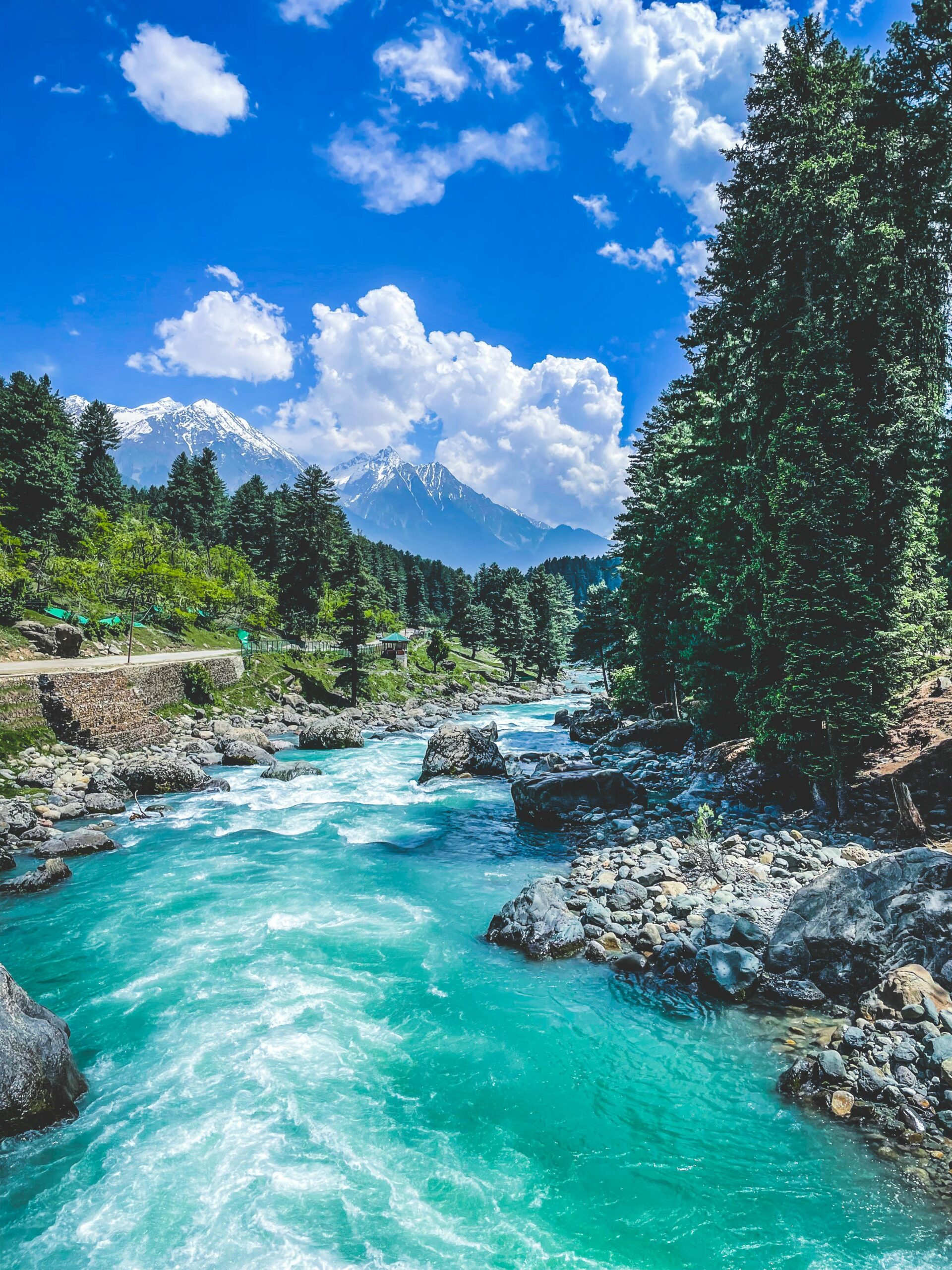 Serene view of a river flowing through Pahalgam's lush mountains, framed by a vibrant blue sky and white clouds.